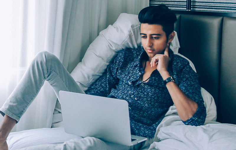 Man reading on a laptop in bed A man reclining on a bed with a laptop, reading thoughtfully
