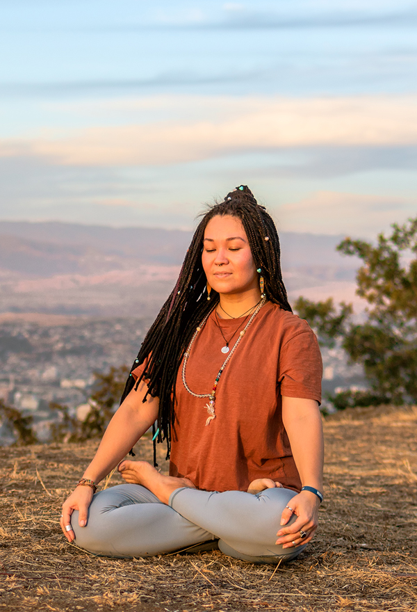 A woman sitting cross-legged in meditation on a hilltop overlooking a valley