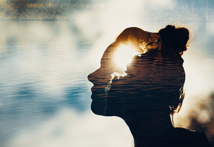 Double-exposure silhouette of a woman's profile filled with a sunset over water, evoking inner landscape and reflection