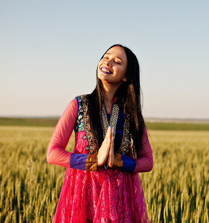 A joyful woman in colorful clothing standing in a green field with eyes closed and hands in prayer mudra, evoking devotion and presence