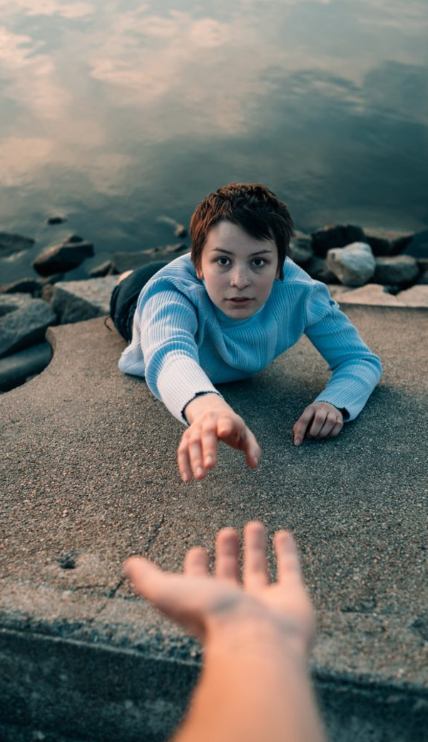 A young woman lying on a ledge above the rocky sea reaching up toward a helping hand, evoking rescue and connection through touch