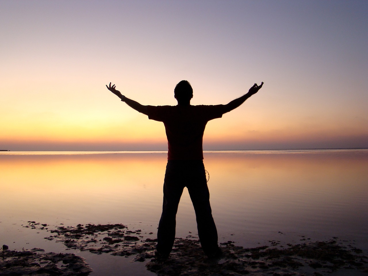 Silhouette of a man facing the sunset with arms open wide, evoking welcoming life and freedom