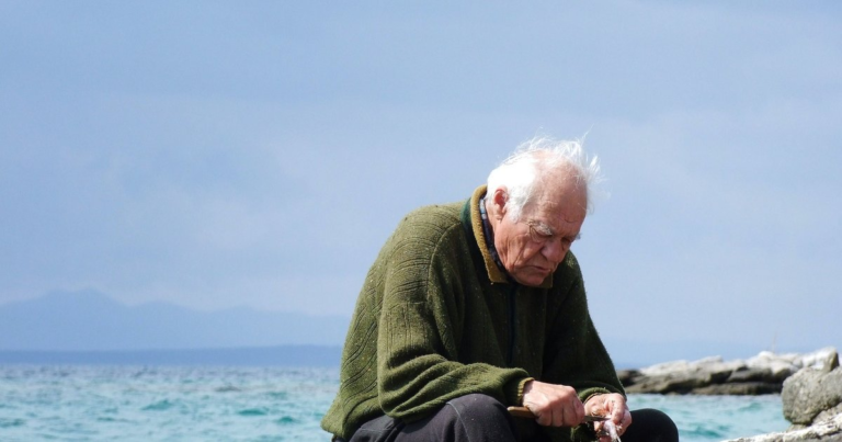An older man in a green sweater sitting alone on rocks beside bright blue water, head bowed over his hands.