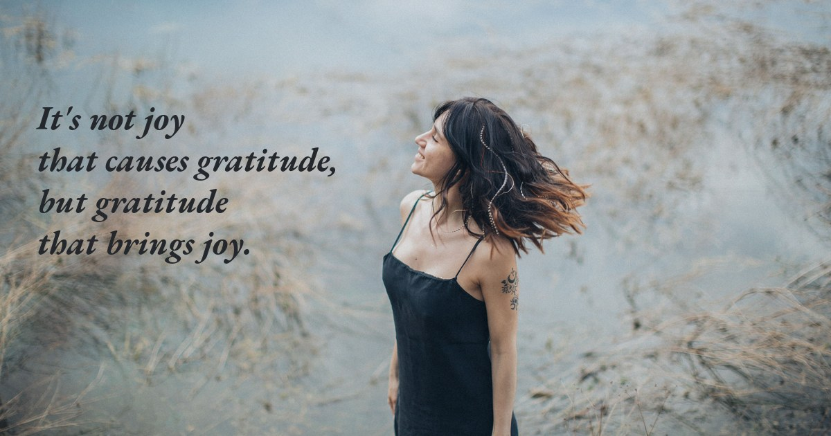 A woman in a black slip dress standing in tall grass at the water's edge, head tipped back with her hair blowing, with the line about gratitude bringing joy set beside her.