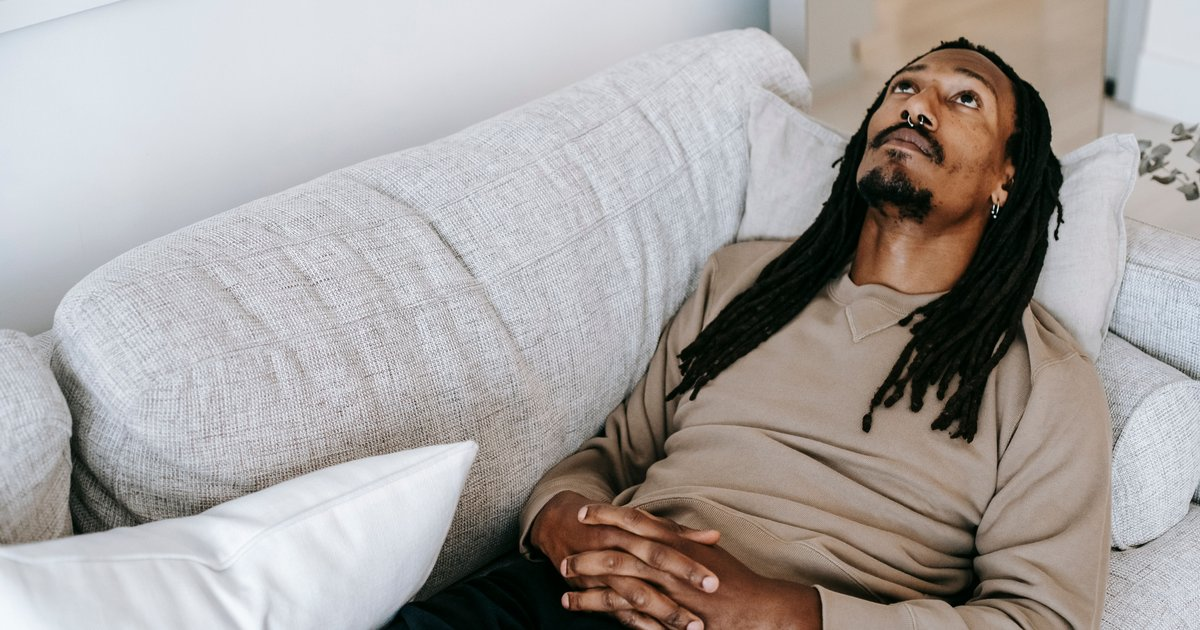 A man lying back on a pale couch with his hands folded on his stomach, looking up at the ceiling.