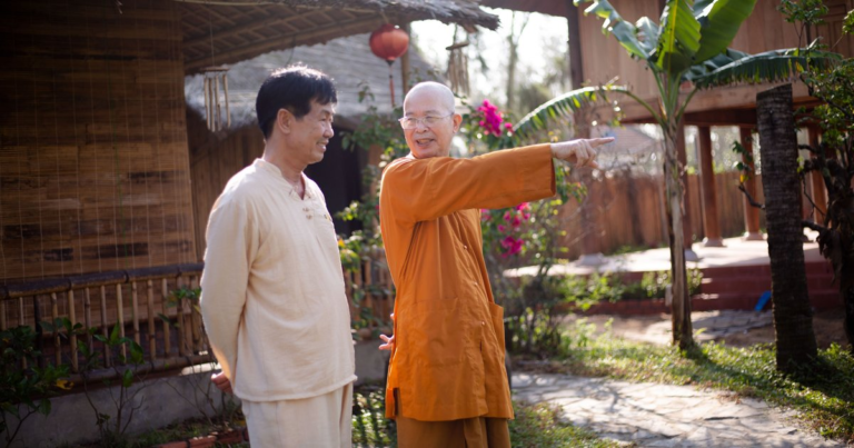 An elder Buddhist monk in orange robes pointing something out to a smiling man in cream linen, the two of them standing together in a sunlit garden of thatched houses and banana palms.