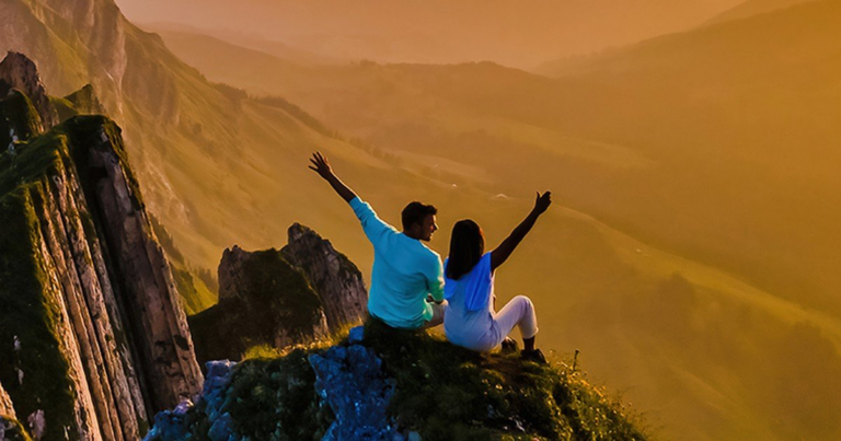 Two people sitting at the edge of a mountain summit at sunrise with arms raised, looking out over a valley of hills.