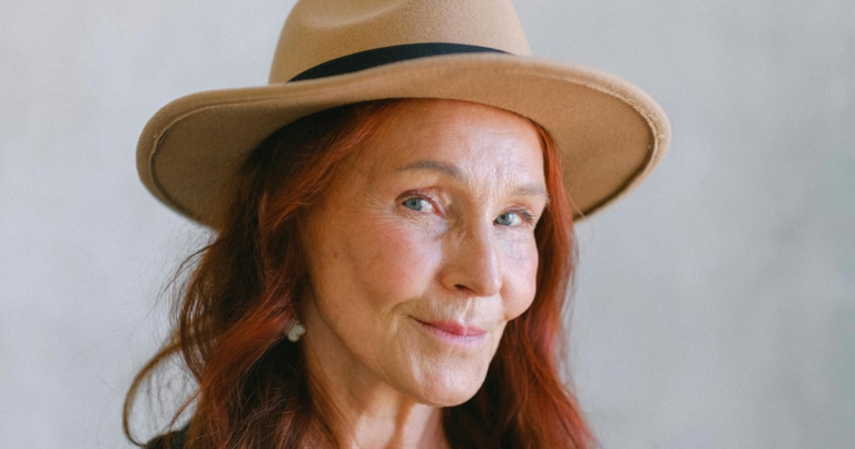 An elder woman with red hair in a wide-brimmed hat, half-smiling at the camera.