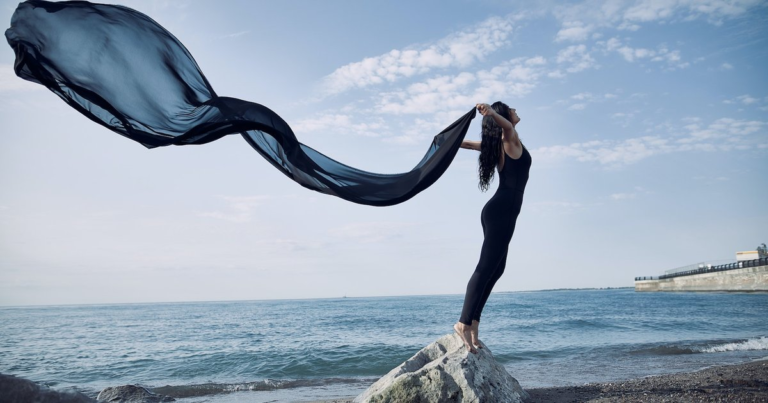 A woman standing barefoot on a rock at the shoreline, arms raised, letting a long dark scarf stream out over the water on the wind.