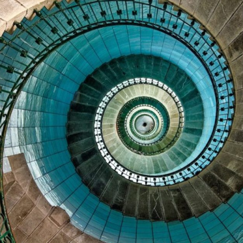 Looking straight down the vortex of a lighthouse spiral staircase, turquoise and stone steps circling inward to a small bright centre.