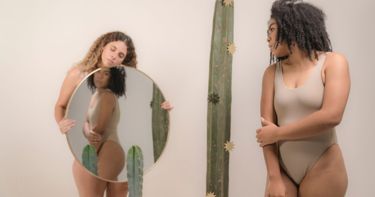 Two women in bodysuits, one holding a round mirror that reflects the other, both looking at their own bodies.
