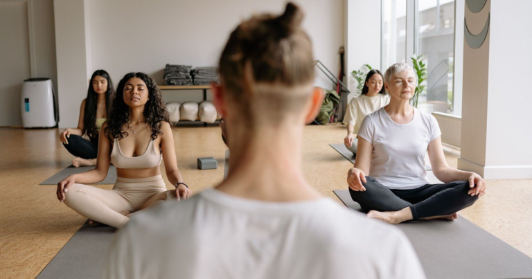 A yoga class seated cross-legged on mats, seen from behind one person's back, with an older white-haired woman meditating among them.