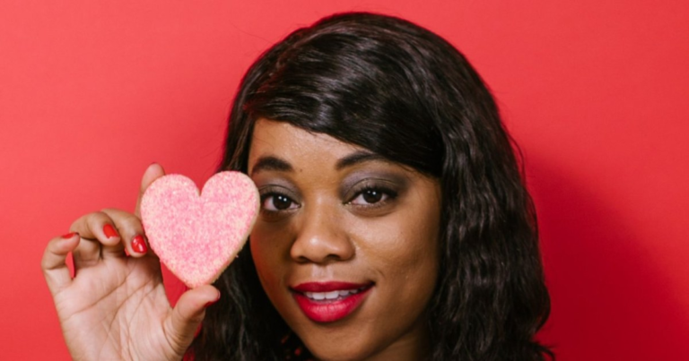 A smiling woman in a blue sweater holds a pink sugar heart cookie up beside her face against a red wall.