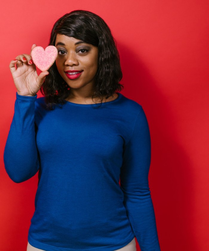A smiling woman in a blue sweater holds a pink sugar heart cookie up beside her face against a red wall.