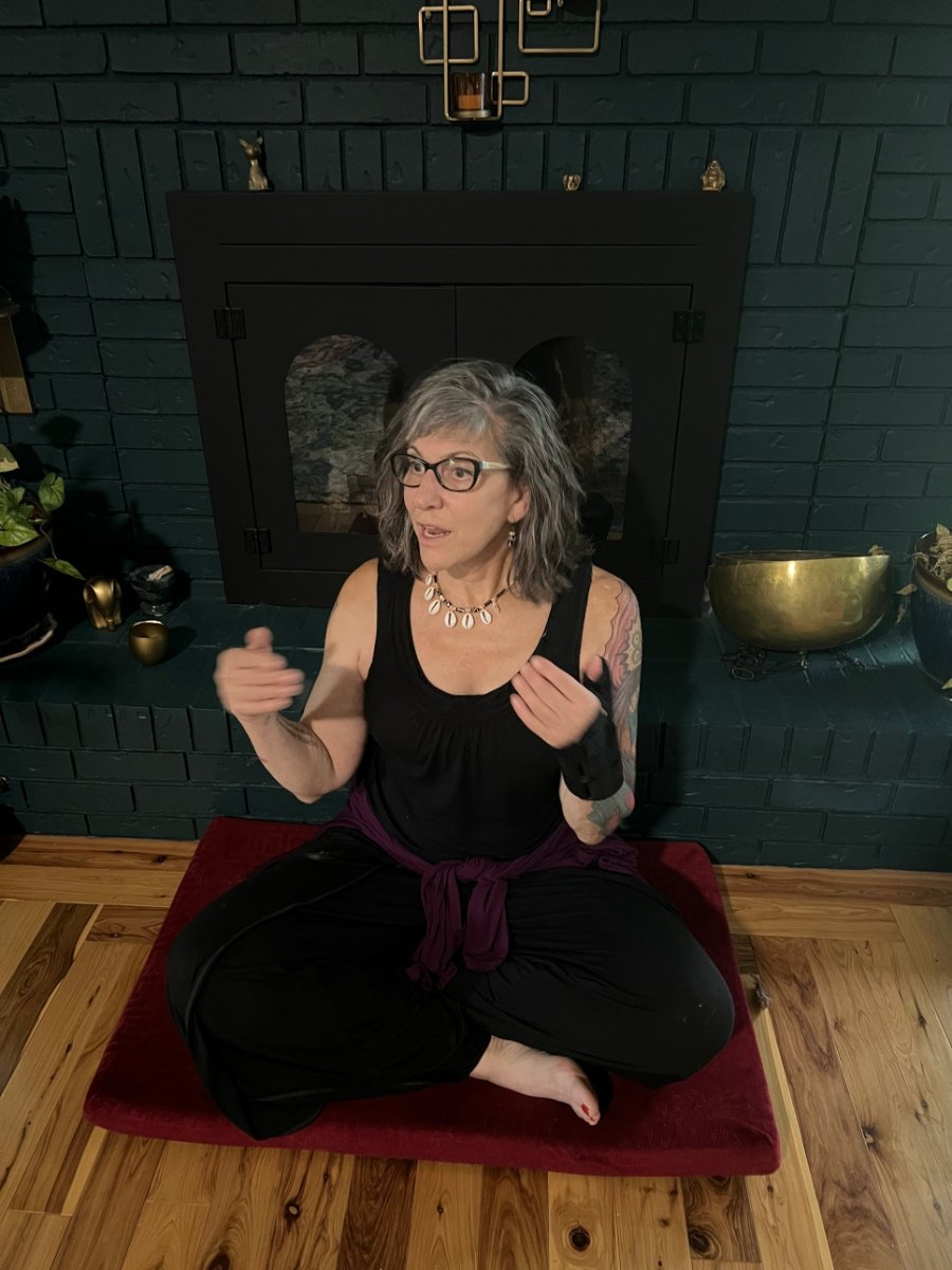 Sabrina Santa Clara sits cross-legged on a red cushion before a dark teal brick hearth, hands lifted as she speaks.