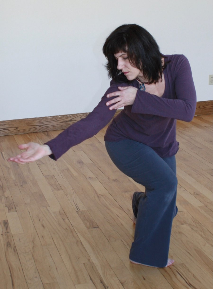 Sabrina Santa Clara, in a plum top, moves low across a wooden floor, one hand on her shoulder and the other arm reaching out.