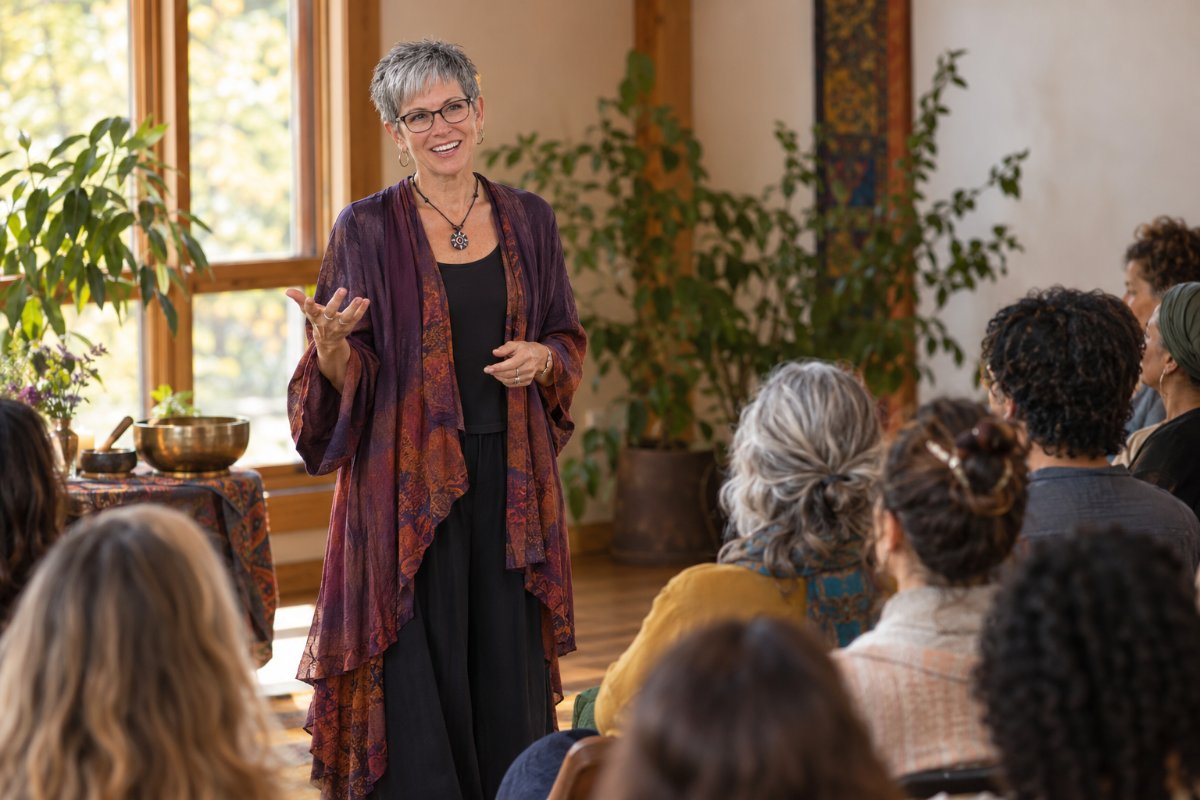 Sabrina Santa Clara, in a flowing plum and rust duster, smiles as she teaches a seated group in a sunlit room with plants and singing bowls.
