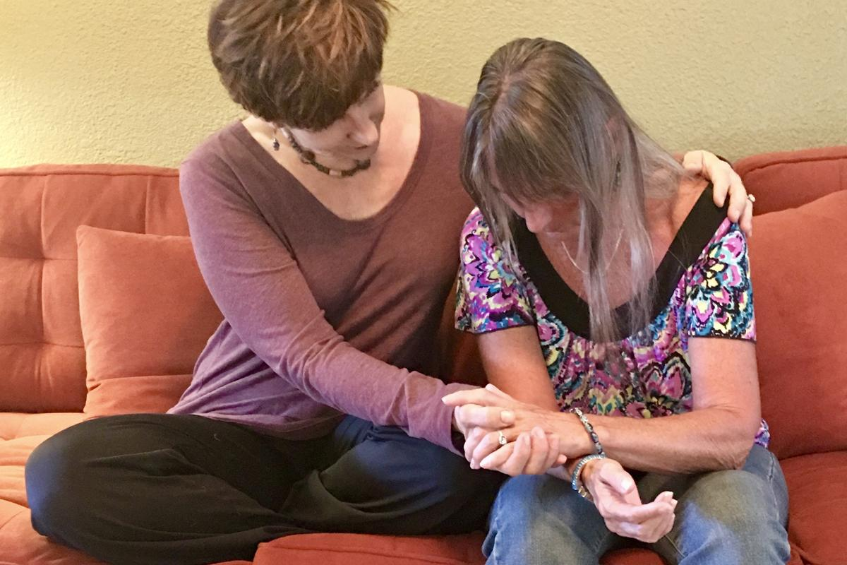 Sabrina Santa Clara sits beside a woman on an orange couch, one arm around her shoulders and a hand cradling hers