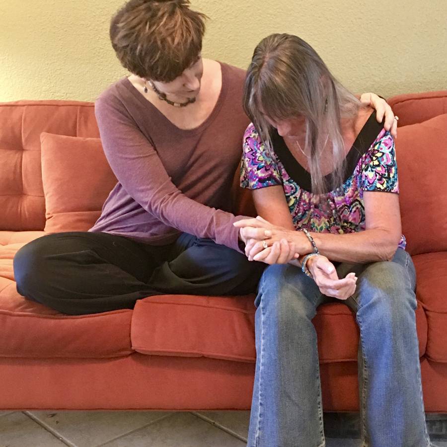 Touch Matters product image Sabrina Santa Clara sits beside a woman on an orange couch, one arm around her shoulders and a hand cradling hers, as the woman bows her head
