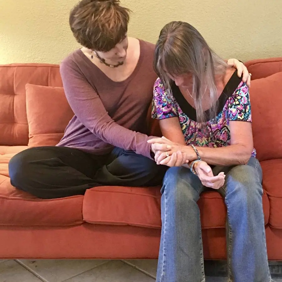 Sabrina Santa Clara sits beside a woman on an orange couch, one arm around her shoulders and a hand cradling hers, as the woman bows her head.