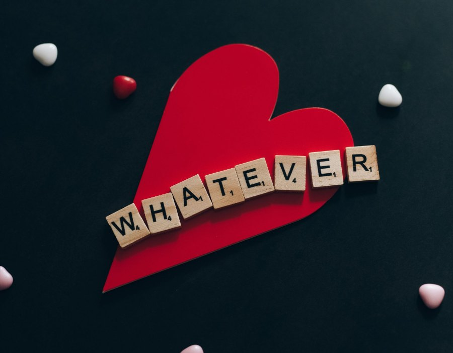 The word WHATEVER spelled in wooden letter tiles across a red paper heart, candy hearts scattered on black.